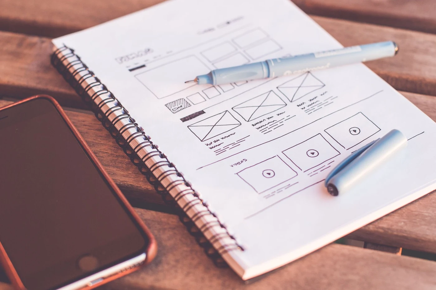 A hand-drawn page layout sketched in a notebook, with a pen and a phone on a wooden table.