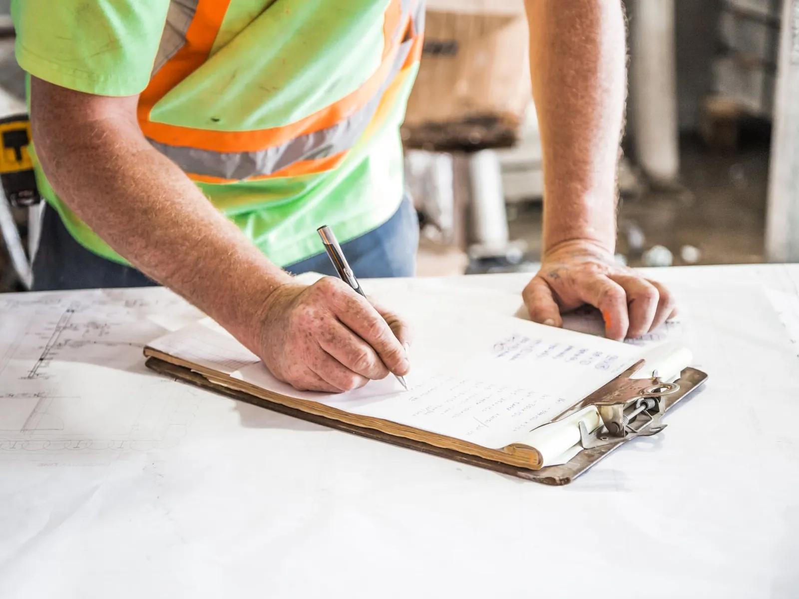 A tradesperson writing notes on a clipboard laid over a set of drawings at a work site.